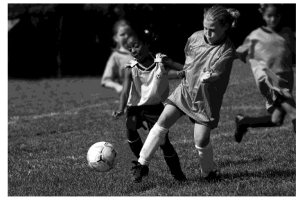 Two young girls playing football, both focused on the ball, with one in an oversized shirt kicking it. Other children and grass in the background.