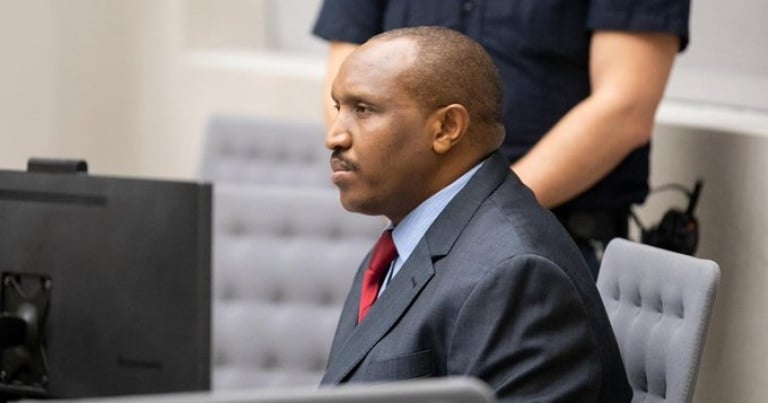 Man in a suit and red tie sitting in court, with a serious expression. A guard stands in the background, partially visible next to a computer.
