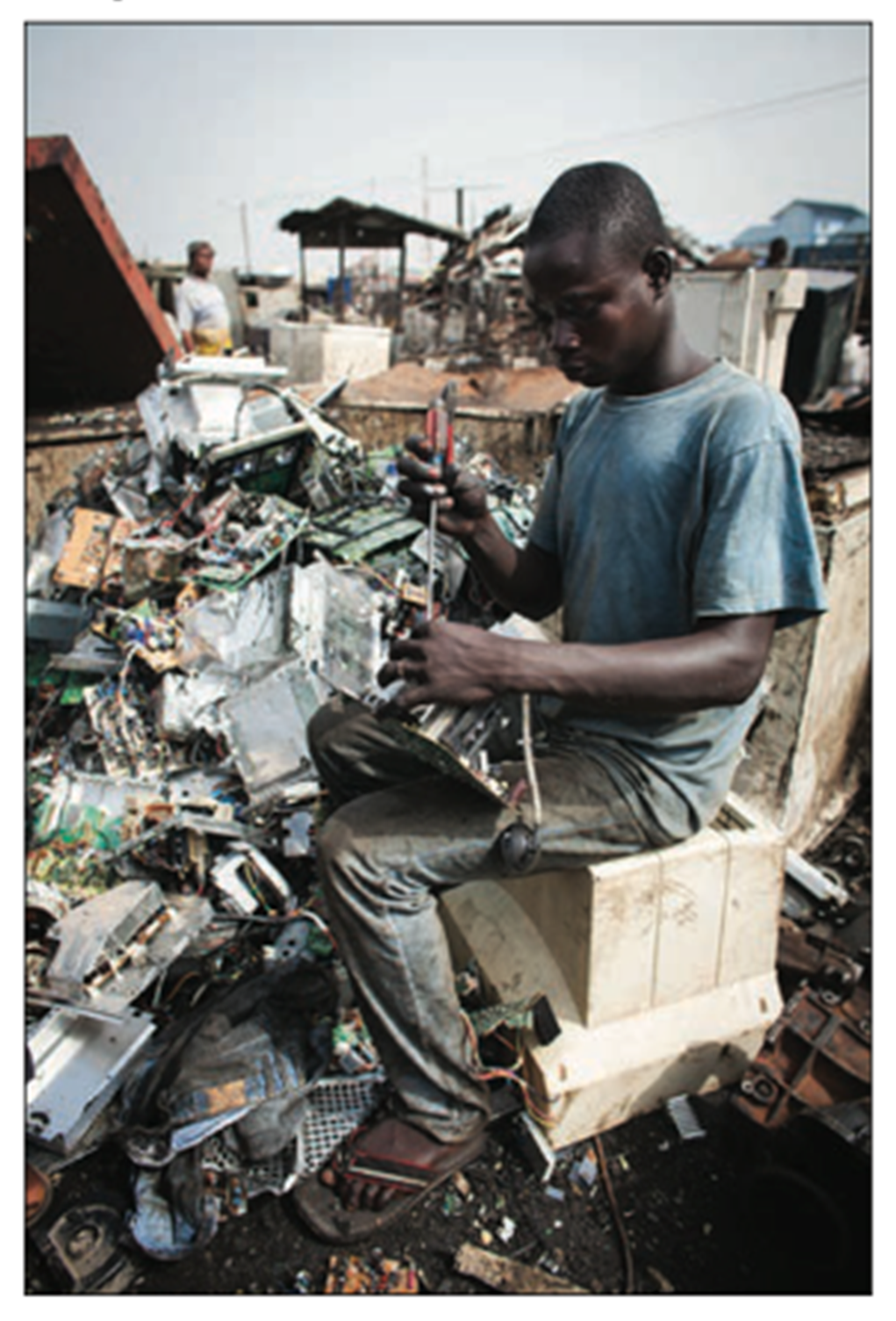 Young man sitting on a plastic crate, using a screwdriver to dismantle electronic waste in a scrapyard, with circuit boards and metallic debris around him.