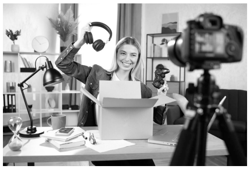 Woman unpacking headphones at a desk with a camera recording, bookshelf and plants in the background; items on the desk include a laptop and mug.
