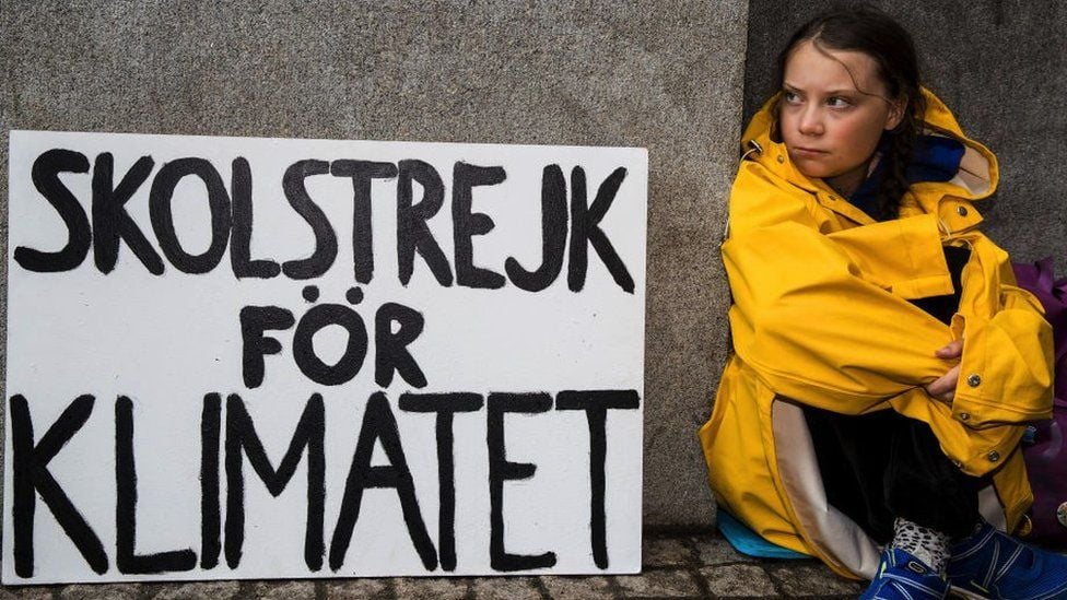 A young person in a yellow raincoat sits beside a sign reading "Skolstrejk för klimatet" against a grey stone wall, conveying a climate protest.