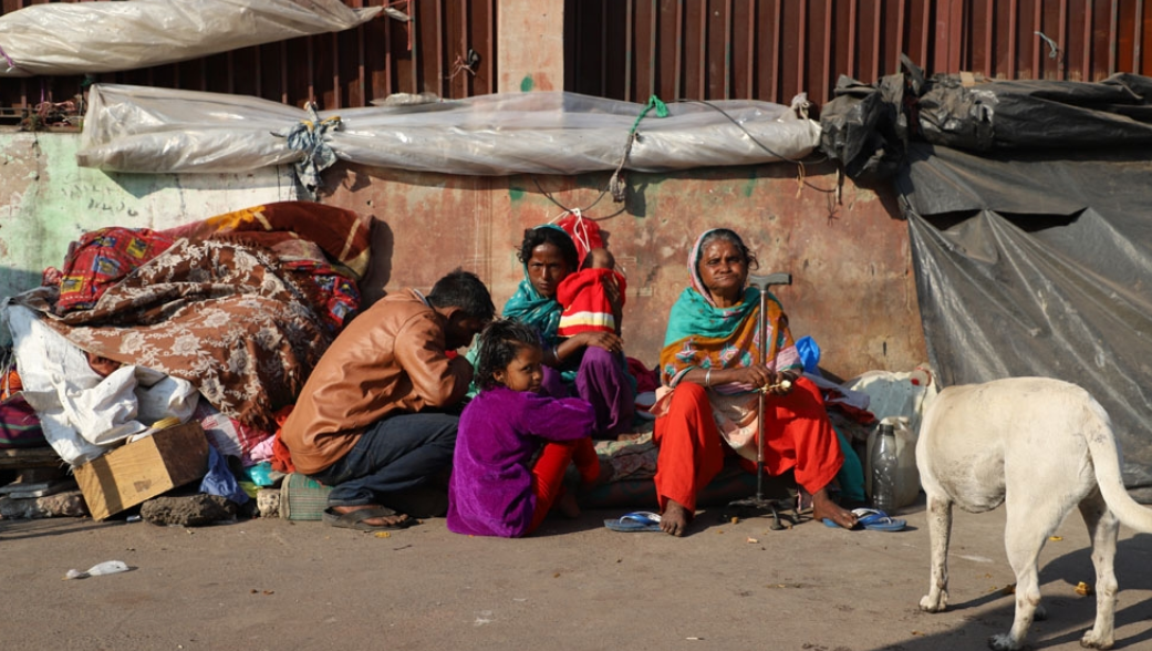 Family sitting on street against a wall; blankets, cardboard, and belongings around them. A dog approaches from the right.