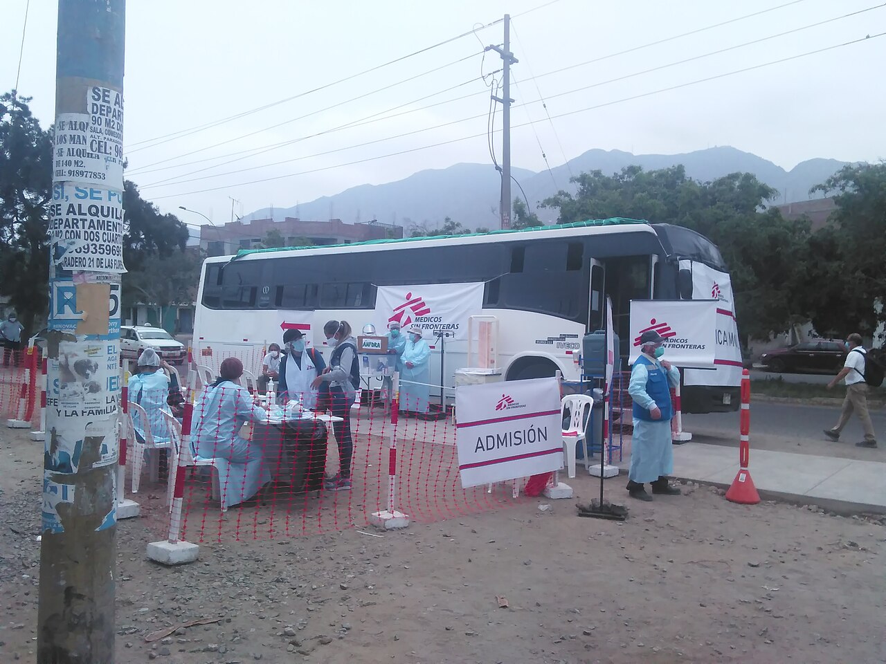 Outdoor COVID-19 testing site with a Médecins Sans Frontières bus. Staff in protective gear assist people, with admission area marked by signs.