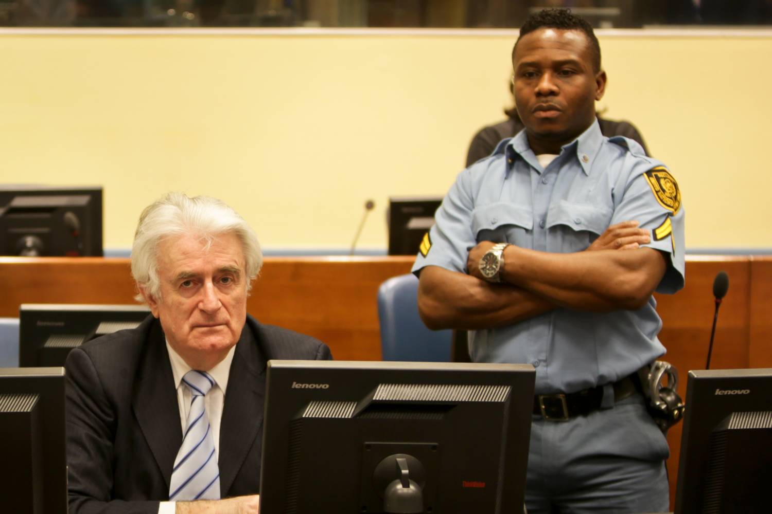 Older man in a suit sits at a desk with computers, watched by a uniformed security officer standing behind with folded arms.