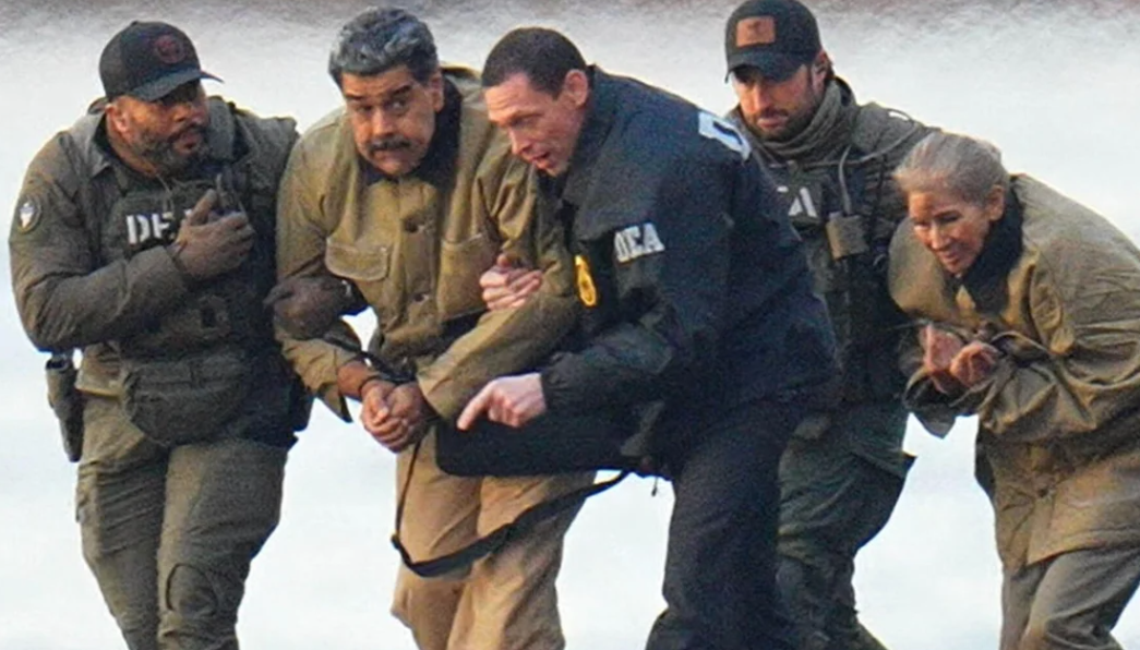 A group of five people, including uniformed officers, assist a handcuffed man in a tan outfit. They appear to be moving quickly outdoors.