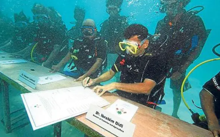 Scuba divers in wetsuits and masks sign documents at an underwater table, with name cards visible, in a unique aquatic setting.
