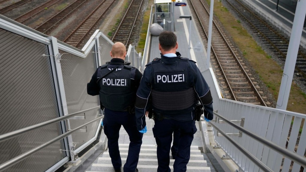 Two police officers in uniform descend metal stairs at a railway station, with tracks visible beside them, carrying blue gloves and equipment.