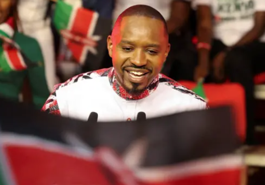 Smiling man wearing a patterned shirt holds a microphone at a public event, with a Kenyan flag in the foreground and people in the background.