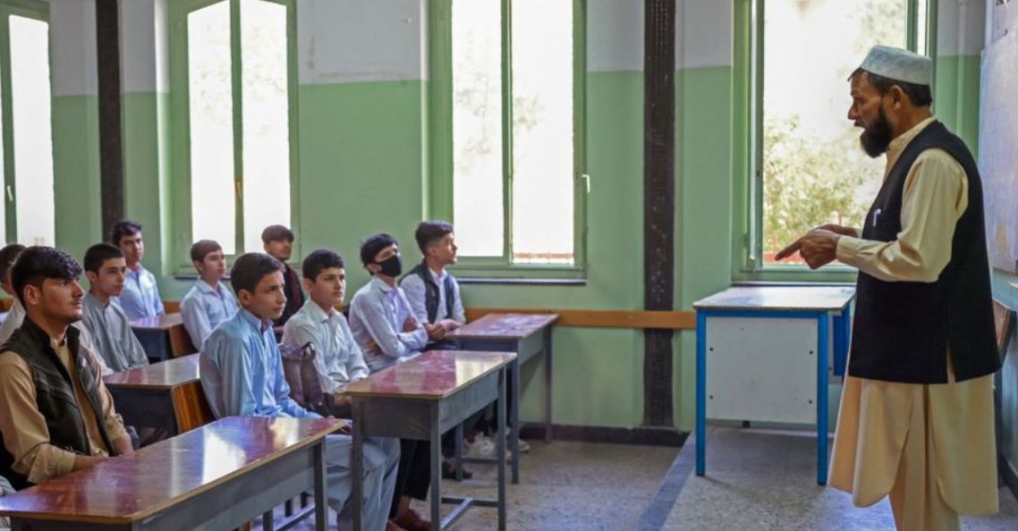 A teacher in traditional attire gestures while standing at the front of a classroom, addressing attentive male students seated at desks.