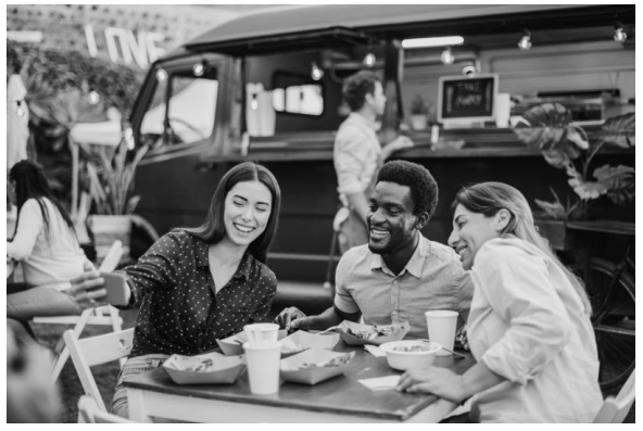 Three people seated at an outdoor table enjoy food and take a selfie. A food truck and other diners are visible in the background.