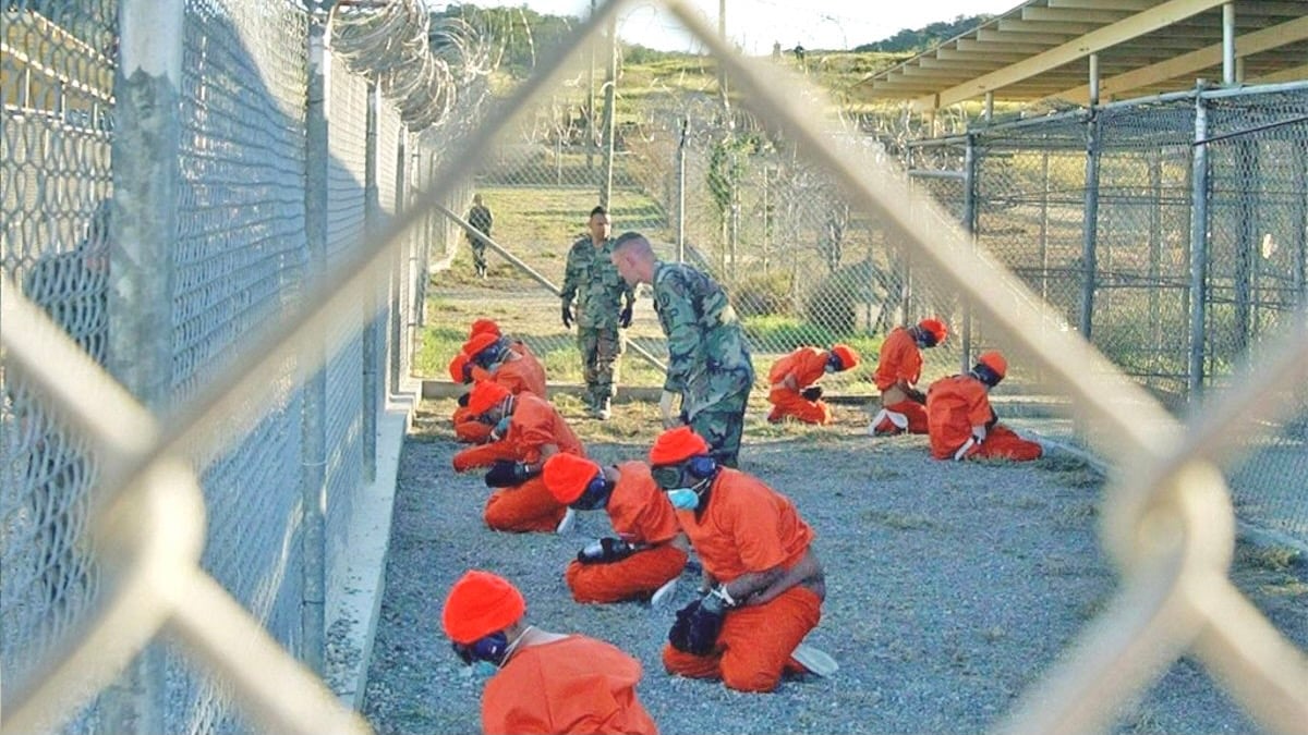 Detainees in orange jumpsuits and red caps kneel in a fenced enclosure, guarded by soldiers in camouflage uniforms, viewed through a chain-link fence.