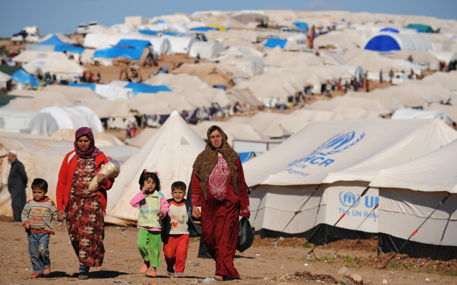Families walking through a refugee camp with numerous tents in the background, including some marked with the UNHCR logo, under a clear blue sky.