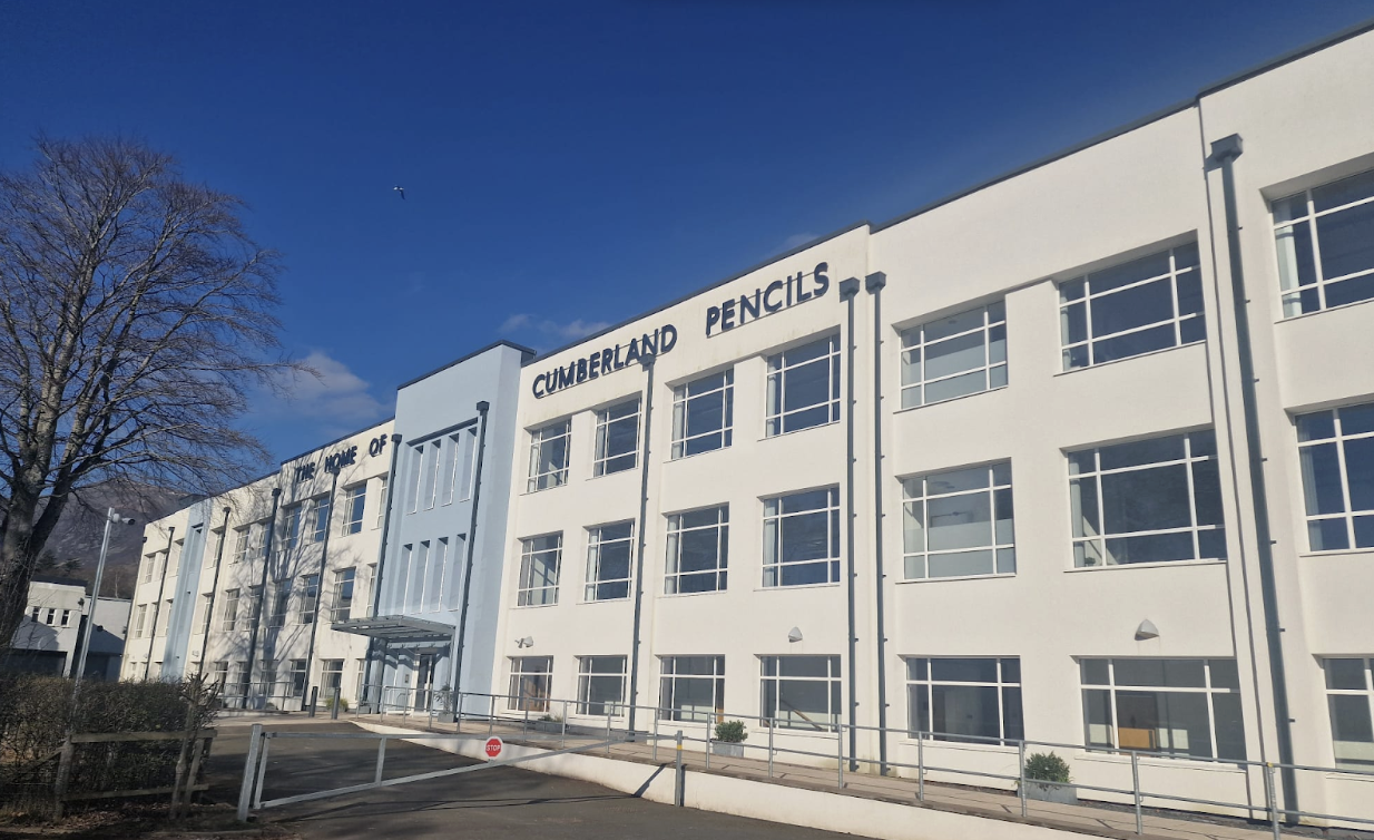 White building with "Cumberland Pencils" sign, large windows, blue accents, set against clear blue sky with bare tree and paved path in foreground.