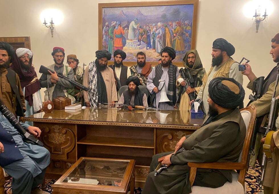 A group of armed men in traditional Afghan clothing surrounds a desk in an office with ornate decor and a large, detailed painting on the wall.
