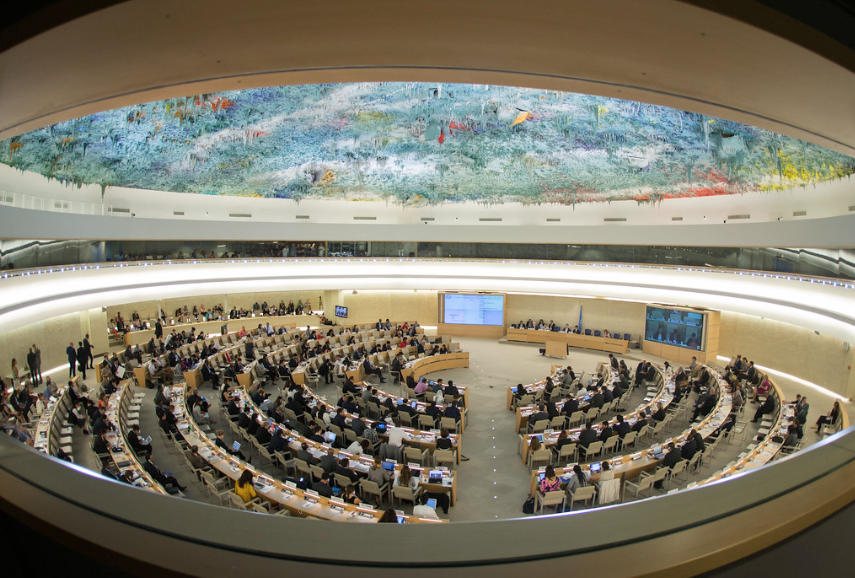 Circular conference room with a vibrant ceiling mural, filled with delegates seated in rows, focusing on large screens and a central speaker's podium.