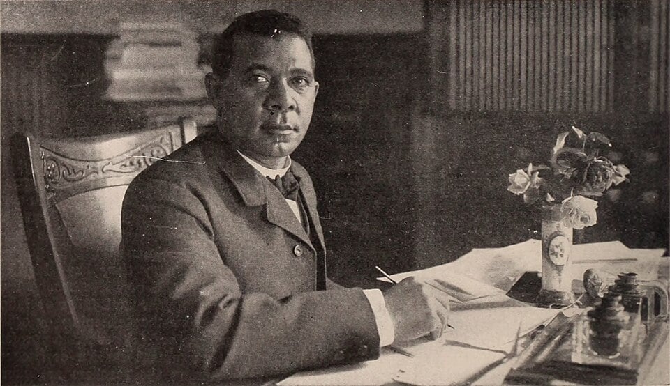 A man in a suit sits at a desk, holding a pen over papers, with a vase of flowers beside him in an office setting.