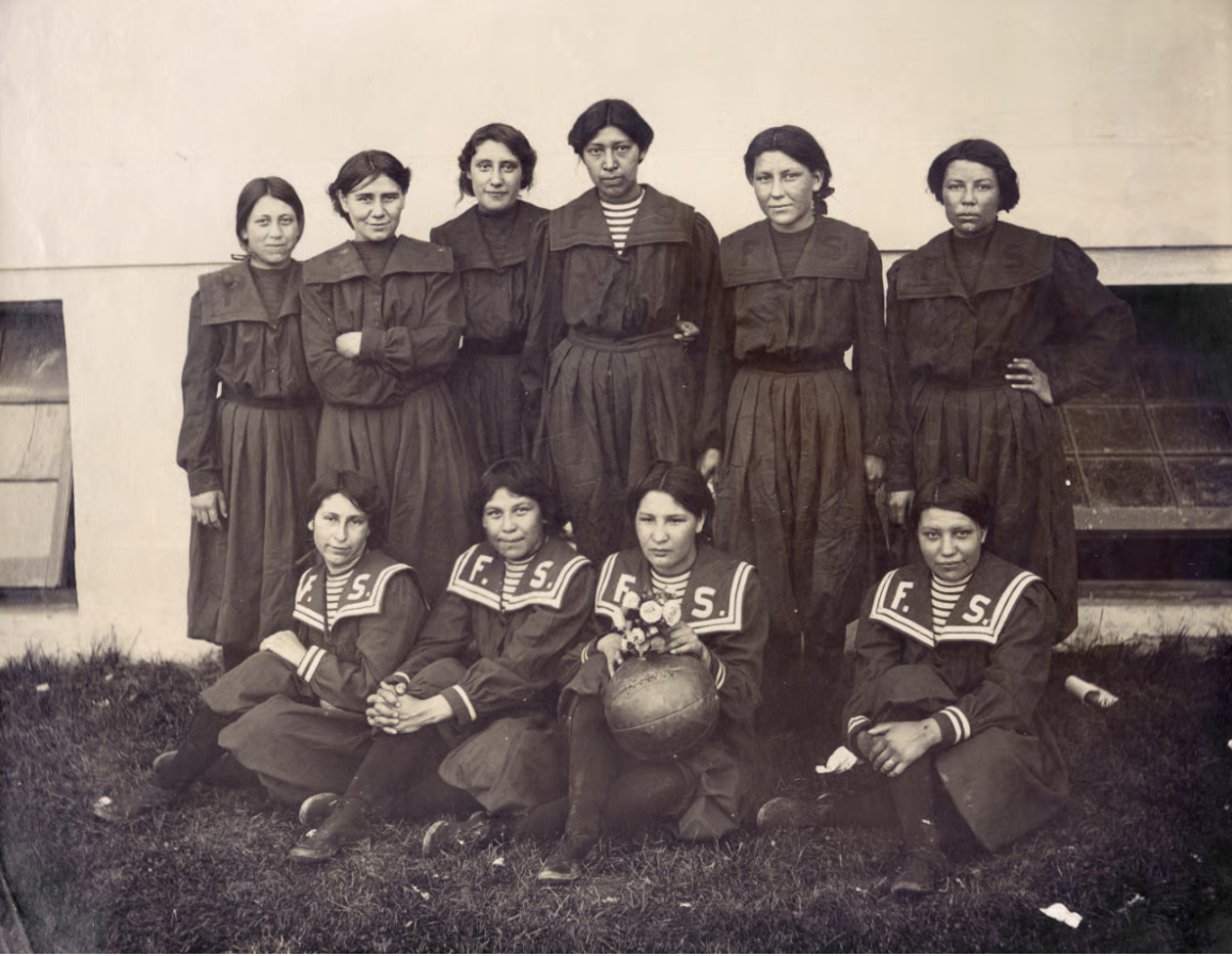 A vintage photograph of ten women, dressed in matching dark uniforms, posed outdoors. The women in front are seated, holding a basketball with "F.S." logo.