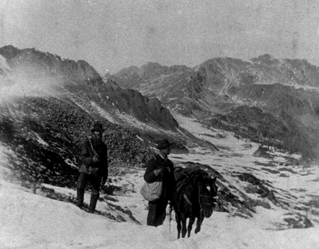 Two men with backpacks and a horse are traversing a snow-covered mountain landscape, with rugged peaks visible in the background.