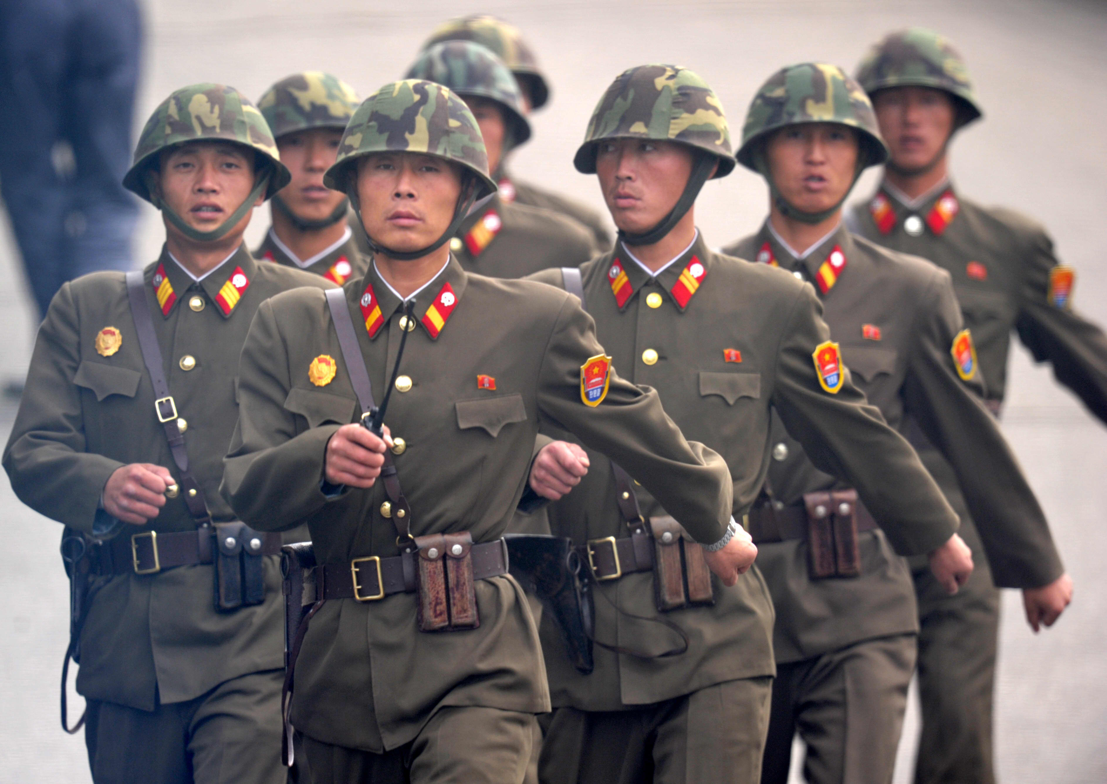 Soldiers in brown uniforms and camouflage helmets march in formation, displaying medals and insignia on their chests and sleeves.