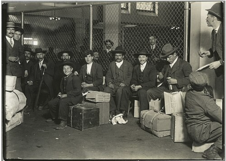A group of men and children, wearing early 20th-century clothing, sit on boxes and luggage in a fenced area, possibly an immigration setting.