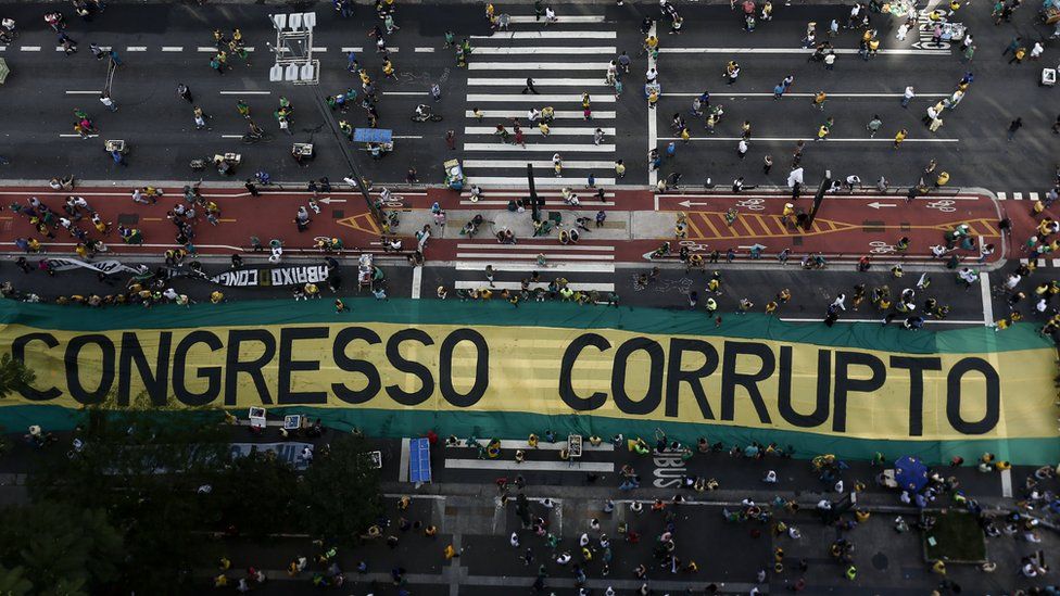 Overhead view of a large protest banner reading "CONGRESSO CORRUPTO" on a street with many people and a zebra crossing.