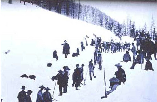 A group of people stand and sit along a snowy mountain path, wearing early 20th-century attire, surrounded by snow-covered trees.