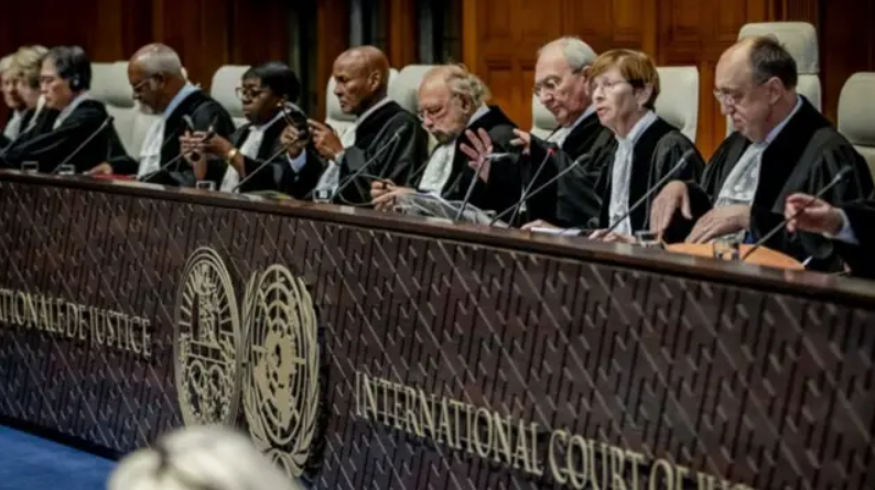 Judges in robes sit at a dais in the International Court of Justice, engaged in discussion, with two emblems visible on the front panel.