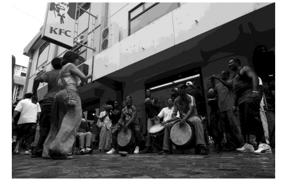 Street musicians play drums as people gather and dance outside a KFC restaurant in a lively urban setting; a festive atmosphere prevails.