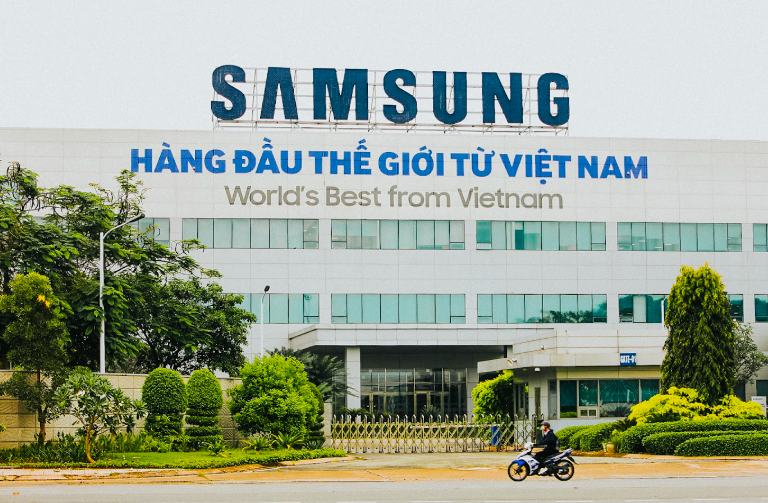 Large building with "Samsung" and "World's Best from Vietnam" signage, surrounded by greenery. A person rides a motorcycle in the foreground.
