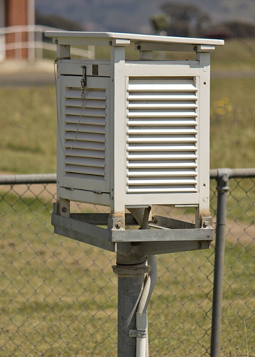 A white Stevenson screen with louvred sides on a metal pole, situated in a grassy area with a chain-link fence in the background.