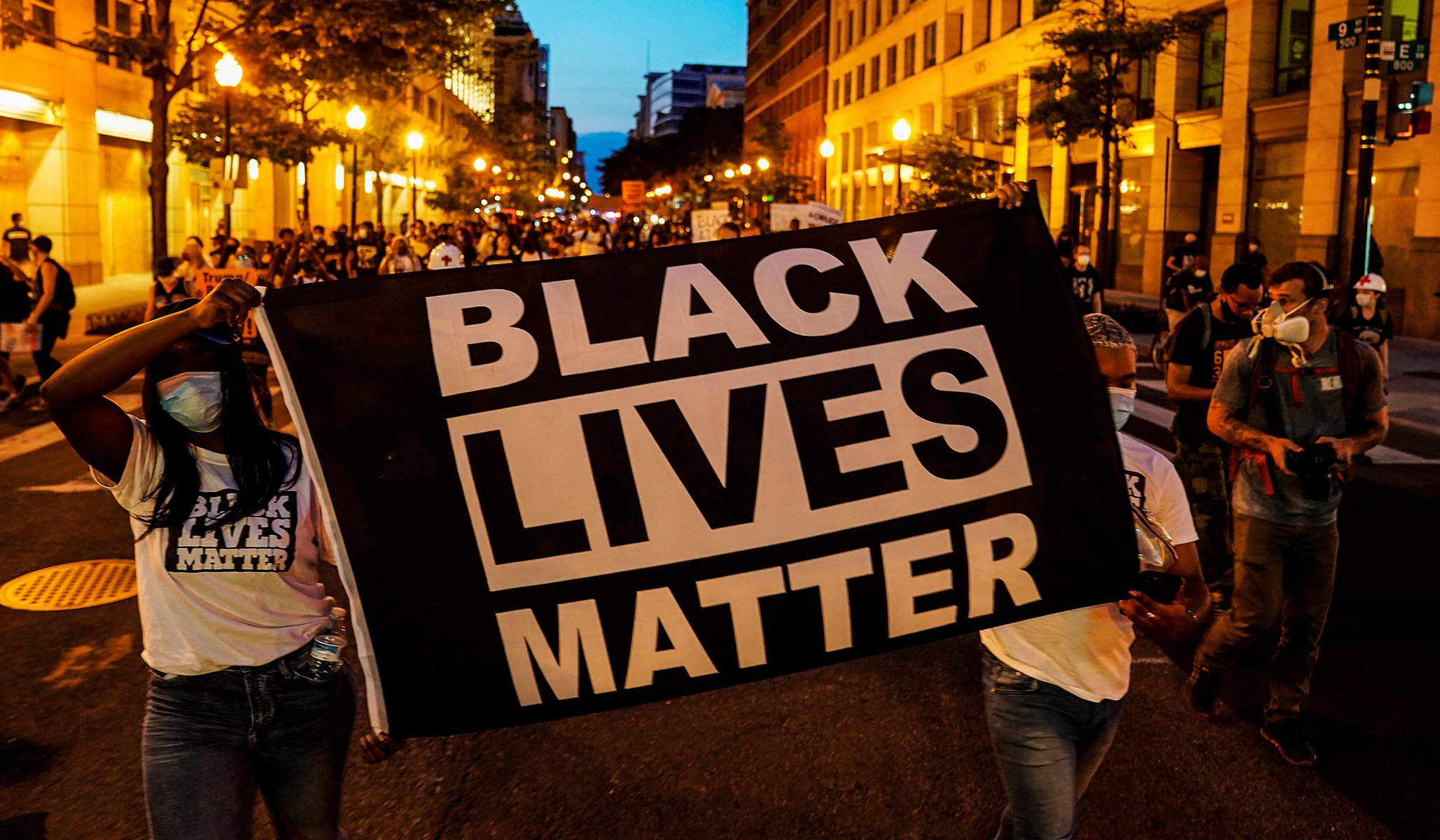 Protesters march down a city street holding a "Black Lives Matter" banner, illuminated by streetlights at dusk, some wearing face masks.