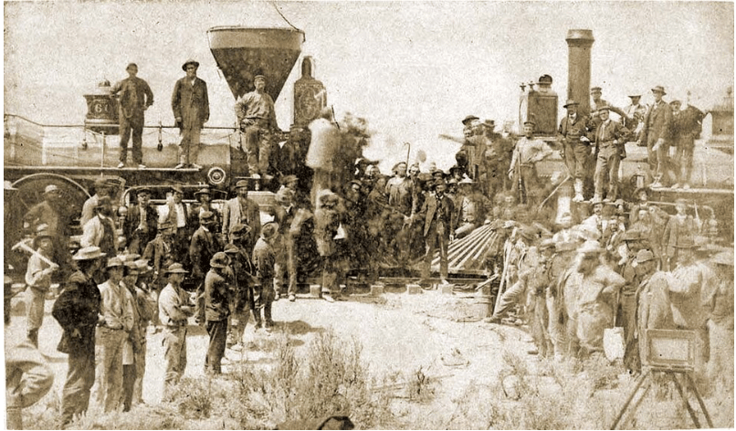 Historic sepia-toned photo of numerous men in 19th-century attire gathered around two steam locomotives on a dusty railway in a celebratory scene.