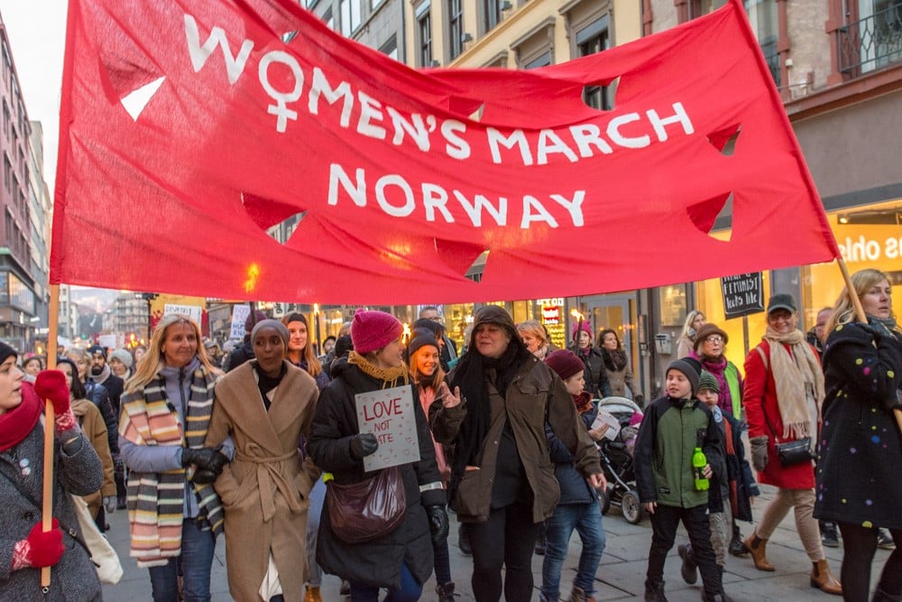 People march in Norway holding a large red banner reading "Women's March Norway." Participants wear winter clothing, and carry signs on a city street.