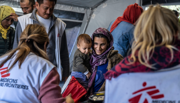 A woman holding a child smiles at aid workers in a tent. The workers wear jackets with the Médecins Sans Frontières logo, assisting people nearby.