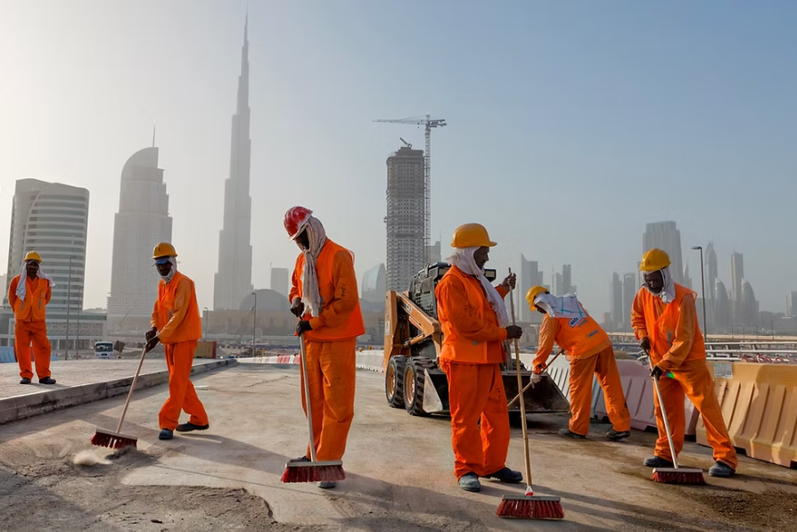 Construction workers in orange uniforms with brooms clean a site in a cityscape with modern skyscrapers, including a tall tower in the background.