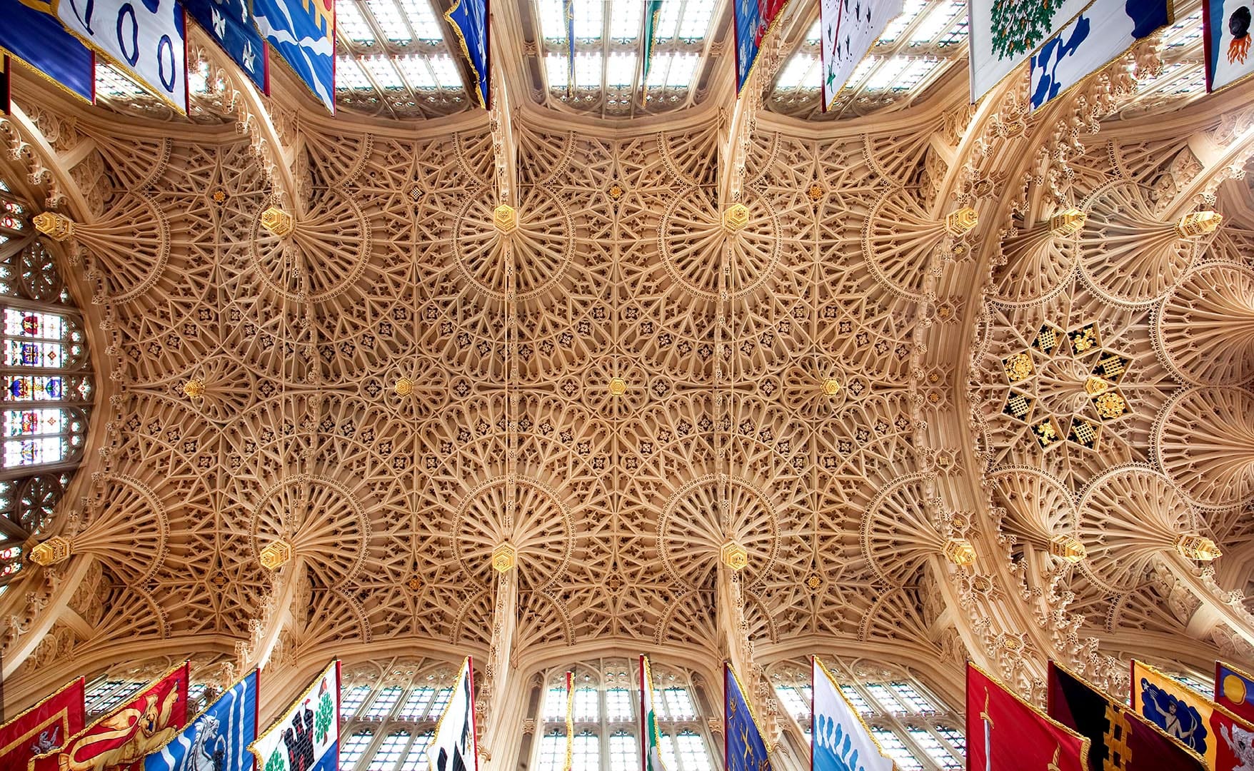 Intricate fan-vaulted ceiling with detailed stonework and colourful heraldic banners beneath stained glass windows in a grand hall.