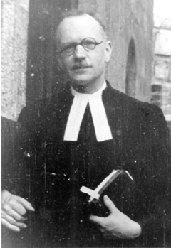 Andre Trocmé in clerical attire with glasses holding a book, stands outdoors beside a stone wall. He wears a clerical collar and looks directly ahead.