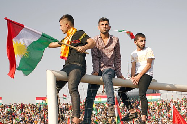 Three men sit on a goalpost holding Kurdish flags, surrounded by a large crowd waving similar flags, under a clear blue sky.
