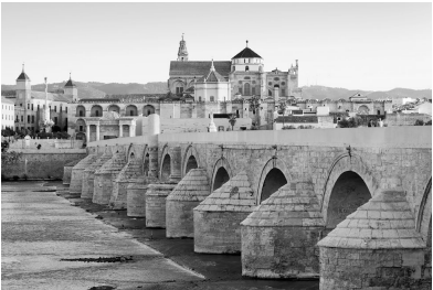 Historic stone bridge over a calm river, with an ancient cityscape and mountainous backdrop under a clear sky.