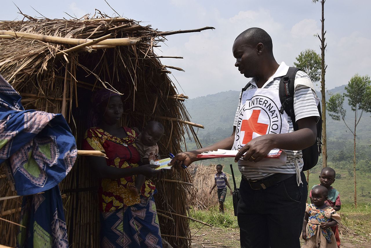 Aid worker with Red Cross vest assisting a woman holding a baby outside a thatched hut in a rural area; two children in the background.