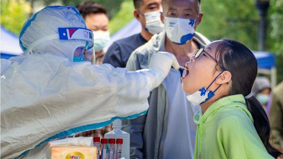 Health worker in protective gear performs a COVID-19 test on a girl in a green hoodie outdoors, with people waiting in the background.