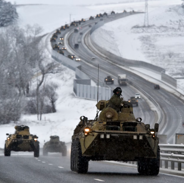 Military vehicles on a snowy highway, leader in foreground, convoy continuing up a winding road, flanked by snow-covered fields and sparse trees.