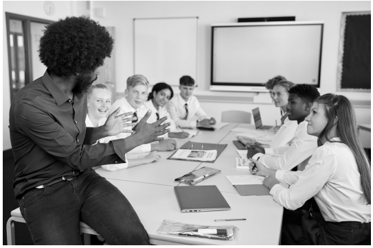 Teacher engaging with diverse students seated around a table in a classroom, discussing enthusiastically with open laptops and notebooks visible.