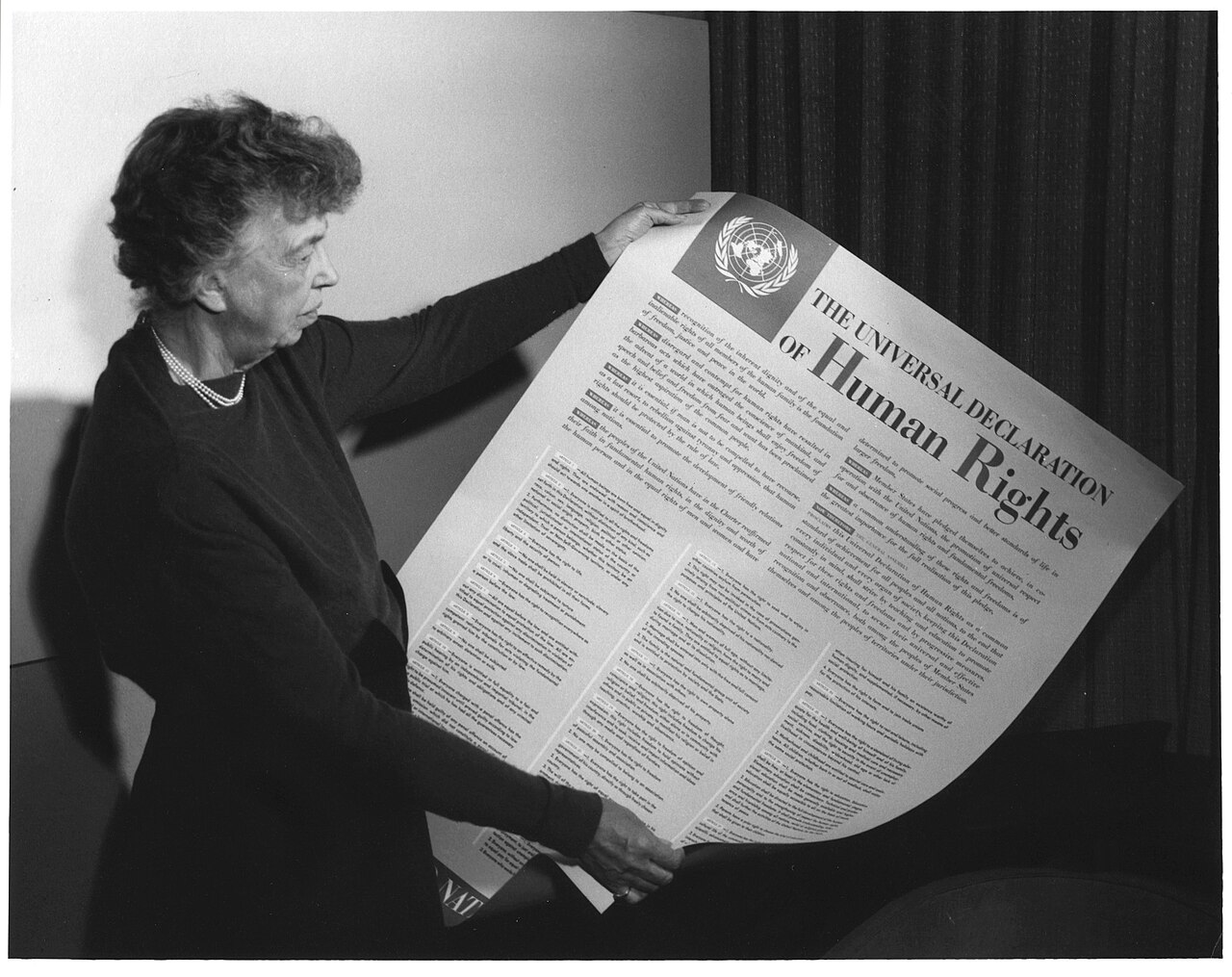 A woman holds and examines the Universal Declaration of Human Rights, a large document with United Nations emblem, in a room with plain and curtain backdrops.