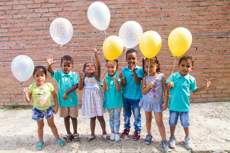 A group of seven young children stand in front of a brick wall, smiling and holding white and yellow balloons on a sunny day.