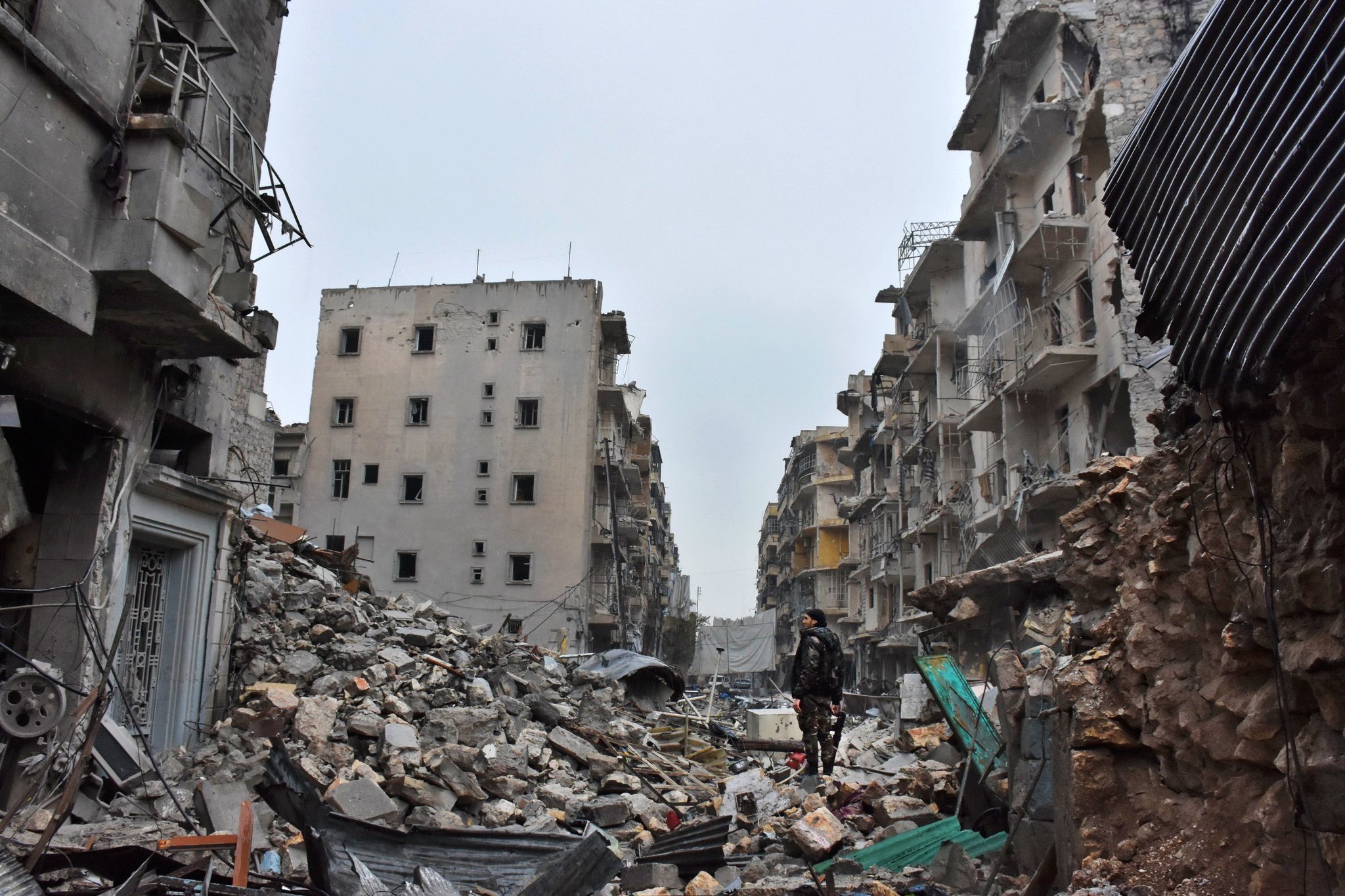 Rubble-strewn street amidst war-damaged buildings, with a lone figure standing among debris, suggesting urban devastation and conflict aftermath.