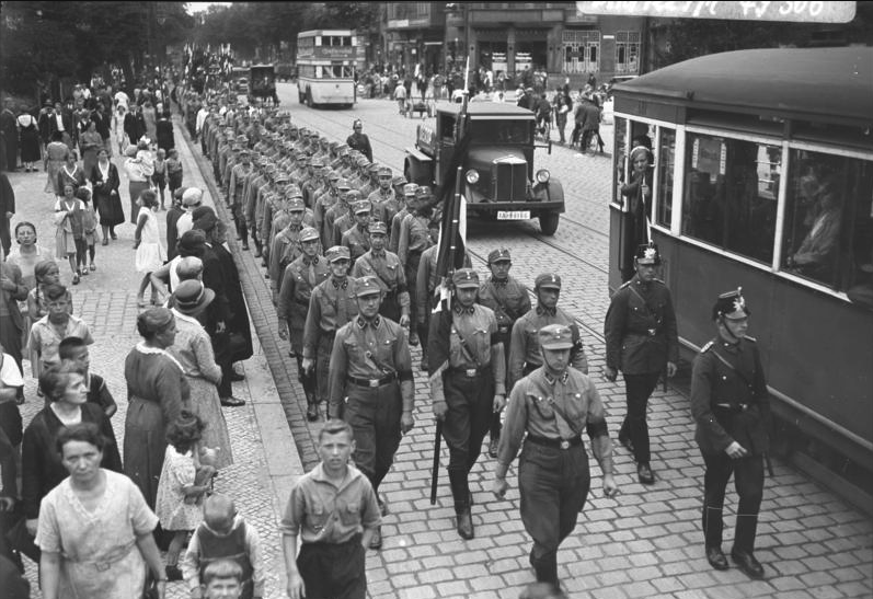 A marching band in uniform parades down a cobblestone street surrounded by onlookers, with vehicles and a tram visible alongside.