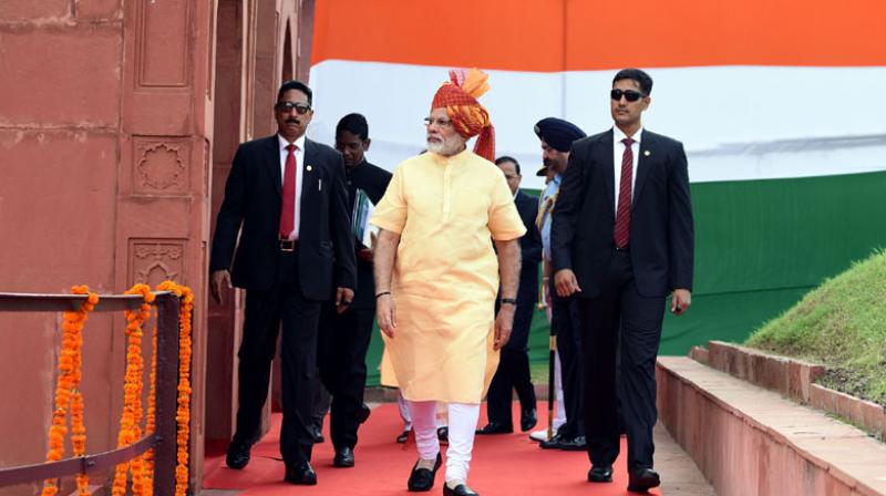 Man in traditional attire with a turban walking on a red carpet, flanked by security personnel, in front of an oversized flag backdrop at a formal event.