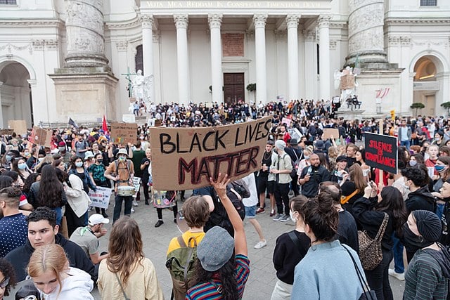 Crowd of people at a protest in front of a historic building, holding signs, including one that reads "Black Lives Matter".