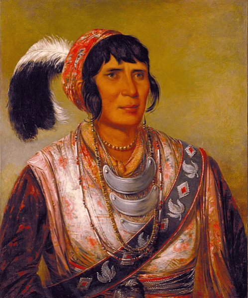 A man in traditional Native American attire with a red headscarf, white feather, layered necklaces, and a decorated sash, set against a plain background.